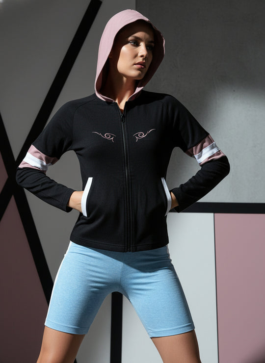 Person wearing a black hoodie with pink hood and white accents, paired with light blue shorts against a geometric background.